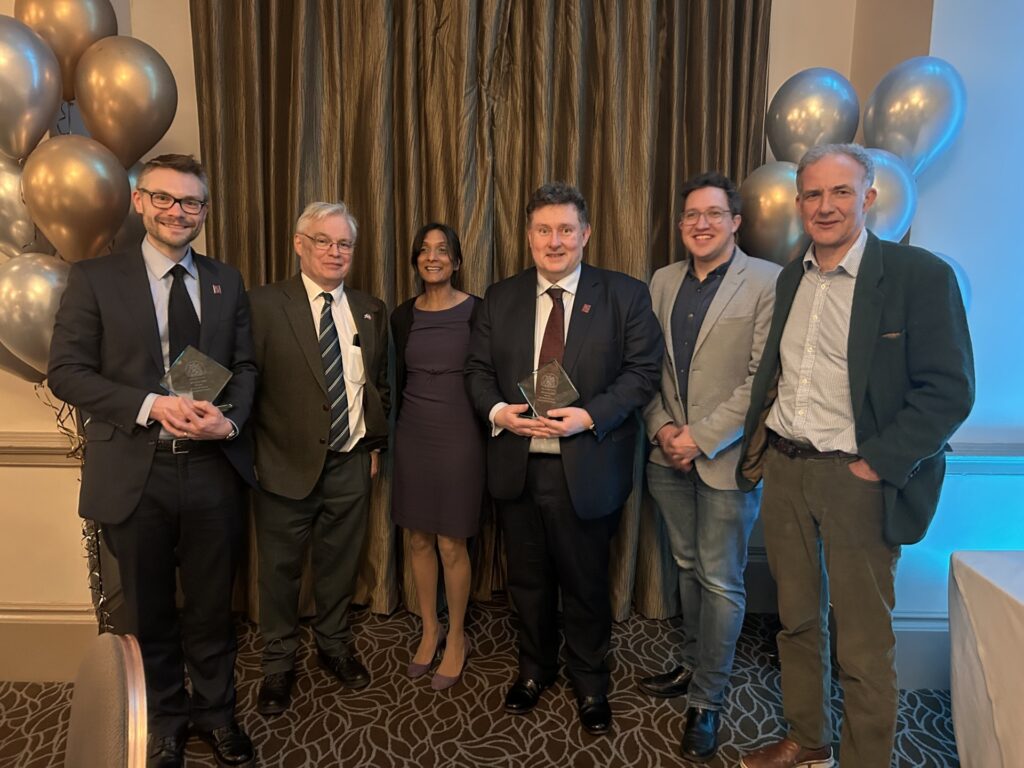 A group of people in smart attire smile at the camera. There are gold and silver balloons in the background.
