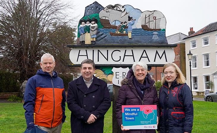 A group of four Wellbeing Champions stand in from of the Hingham village sign holding a sign which says "We are a mindful town".