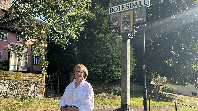 Janet Russell sits below the Botesdale village sign. She is smiling at the camera.