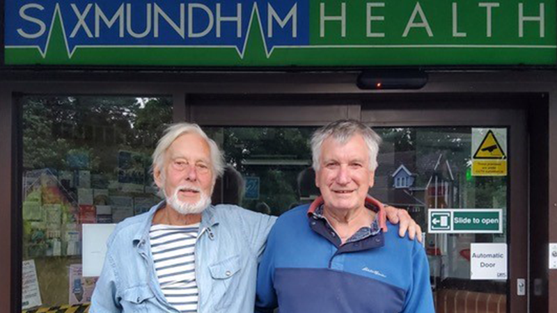 Dr Hilary Graham and Mike Golding stand with their arms around each others' shoulders and smile at the camera. They are outside the Saxmundham Health Centre.