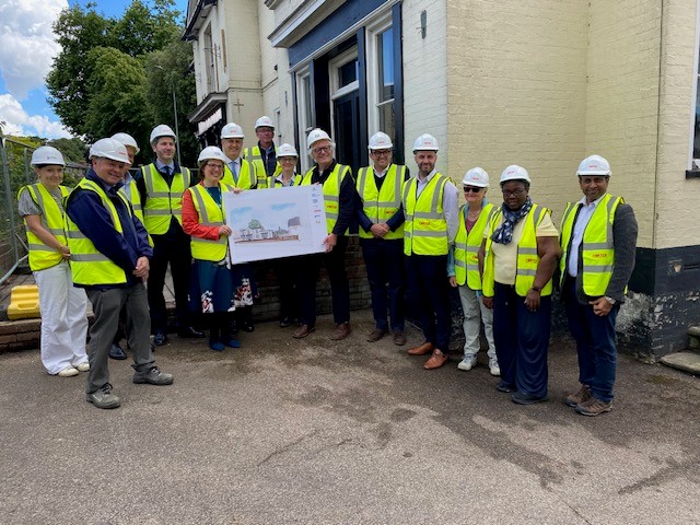 14 people in high-vis vests and hard hats are gathered outside the former Inkerman Pub. Three of them are holding a board with an artist's impression of the new medical practice.