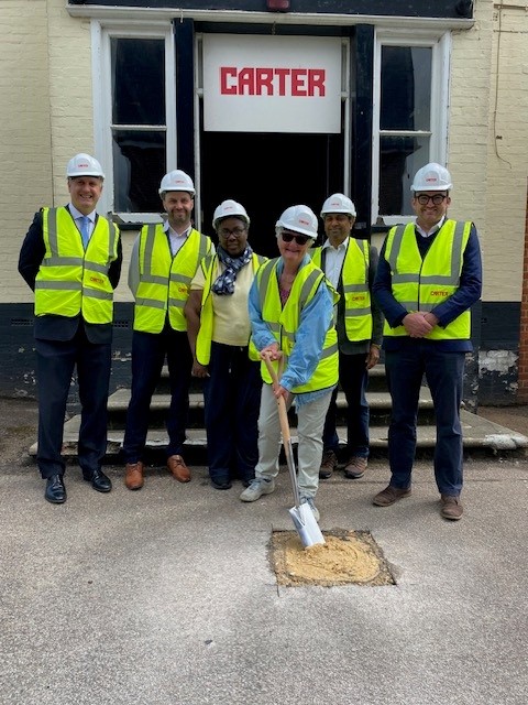 Six members of the project group in hard hats and high-vis vests outside the former Inkerman Pub. Chris - a member of the Patient Participation Group - is holding a spade which has broken the ground marking the start of construction.