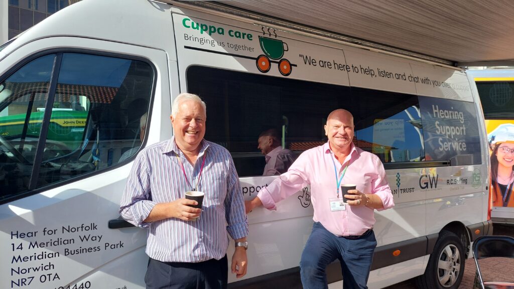 Two representatives from Hear for Norfolk standing in front of their Hearing Support Service van holding hot drinks