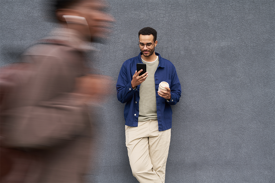 Young man leaning against a wall. He is holding a takeaway coffee cup and looking at his phone screen.
