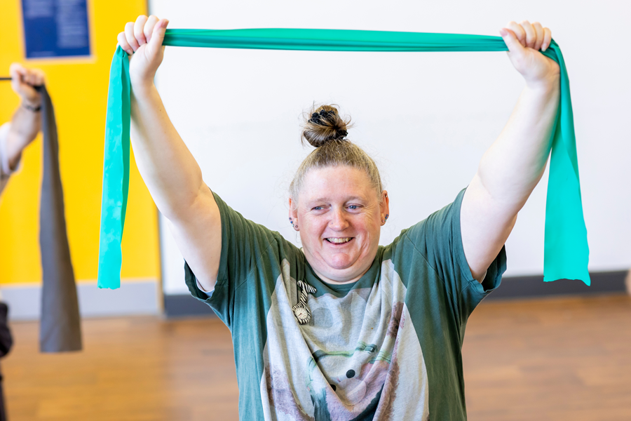 A woman holds her hands above her head and is stretching a blue resistance exercise band.