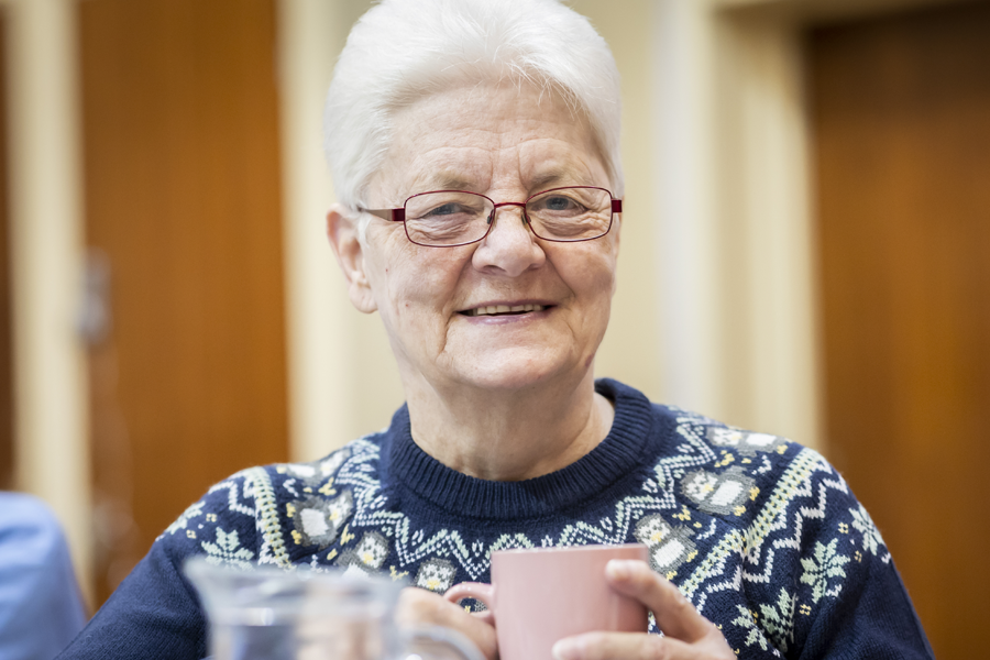 Older woman smiles at the camera. She is wearing a blue festive knitted jumper and holding a coffee cup.