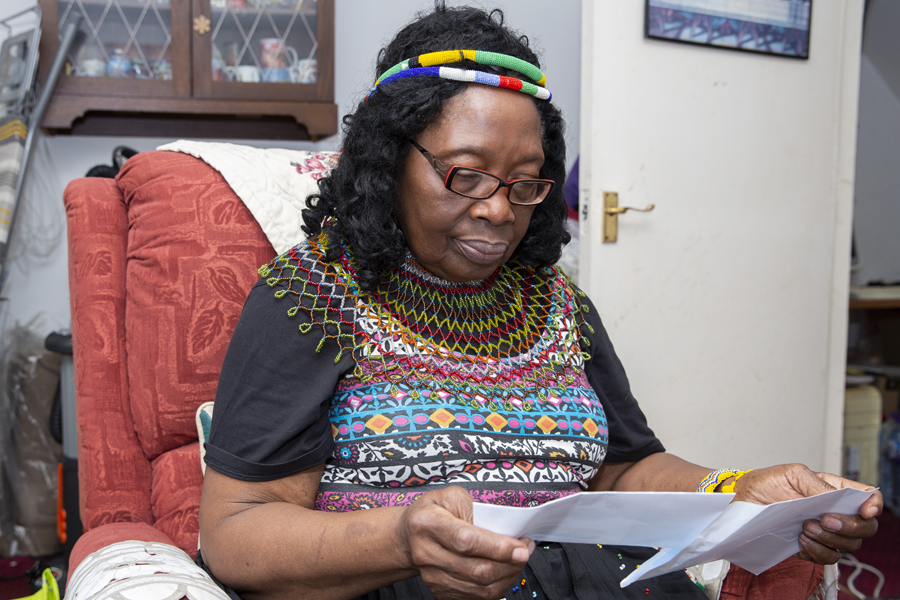 Older woman reading a document. She is sitting on a red armchair in a residential living room.