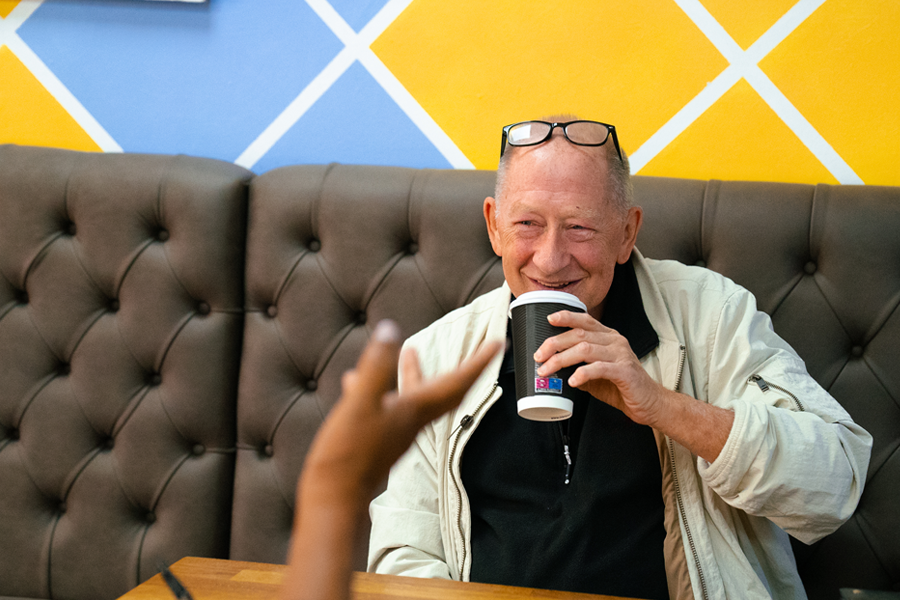 Older man is raising a coffee cup to his lips and smiling at his companion who is behind the camera