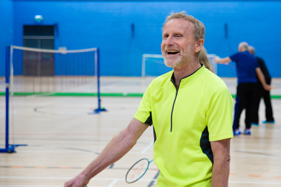 Older man in a bright yellow sports top plays badminton in a sports hall