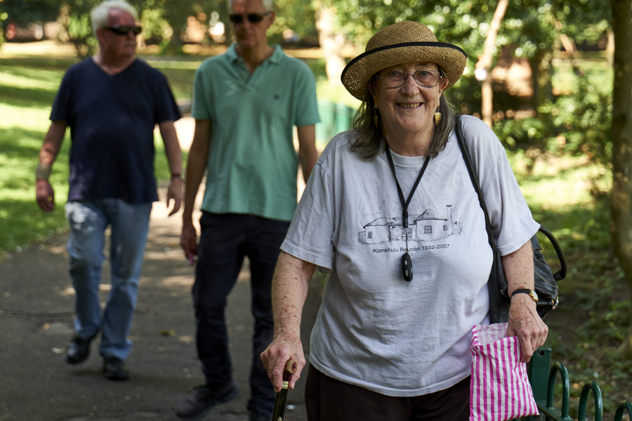 A woman using a walking stick smiles at the camera. She is walking through a park on a sunny day.