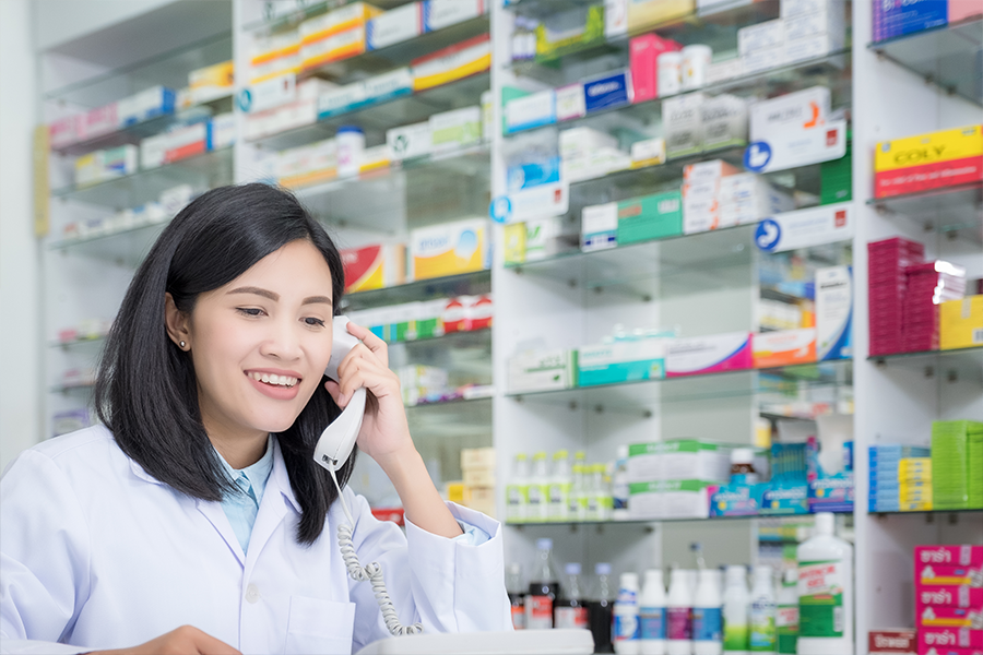 Young pharmacist speaking on the phone. Behind her are shelves stacked with various medications.